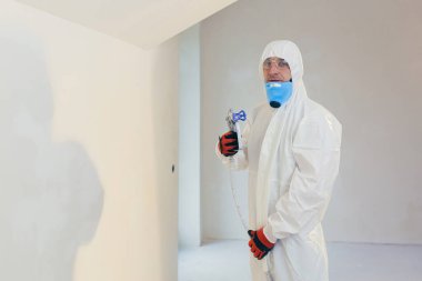 A worker painter paints the surface of the wall with a special spray. Male worker in protective overalls kid surface in respirator. An experienced repairman looks into the camera