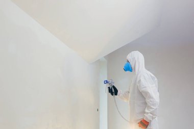 A worker painter paints the surface of the wall with a special spray. Male worker in protective overalls kid surface in respirator. Experienced repairman