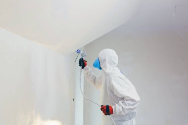 A worker painter paints the surface of the wall with a special spray. Male worker in protective overalls kid surface in respirator. Experienced repairman