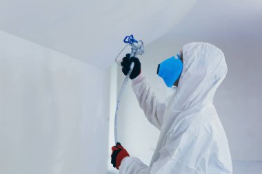 A worker painter paints the surface of the wall with a special spray. Male worker in protective overalls kid surface in respirator. Experienced repairman