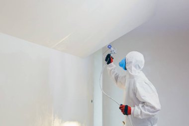 A worker painter paints the surface of the wall with a special spray. Male worker in protective overalls kid surface in respirator. Experienced repairman