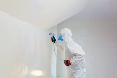 A worker painter paints the surface of the wall with a special spray. Male worker in protective overalls kid surface in respirator. Experienced repairman
