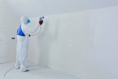 A worker painter paints the surface of the wall with a special spray. Male worker in protective overalls kid surface in respirator. Experienced repairman
