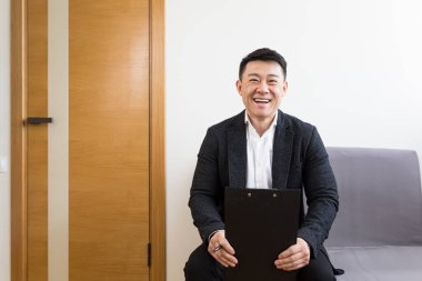 Asyalı erkek işadamının iyi haberleri var. Modern bir iş merkezinin resepsiyonunda röportaj için bekleme odasında seviniyor.