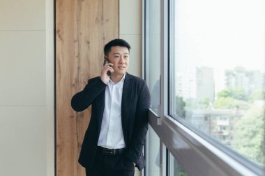 Mutlu ve gülümseyen Asyalı işadamı modern bir ofis merkezinde telefonla konuşuyor, iyi haberleri arkadaşlarıyla paylaşıyor.