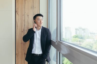 ciddi patron Asyalı iş adamı modern bir ofis merkezinde telefonla konuşuyor, iş arkadaşlarına iş görüşmesi yapıyor.