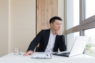 Ofiste baş ağrısı çeken genç Asyalı ofis çalışanı suyla birlikte ilaç içiyor. İçerideki bilgisayarın başında ağır ağrısı olan hasta bir adam ilacı iş yerinde kullanıyor.