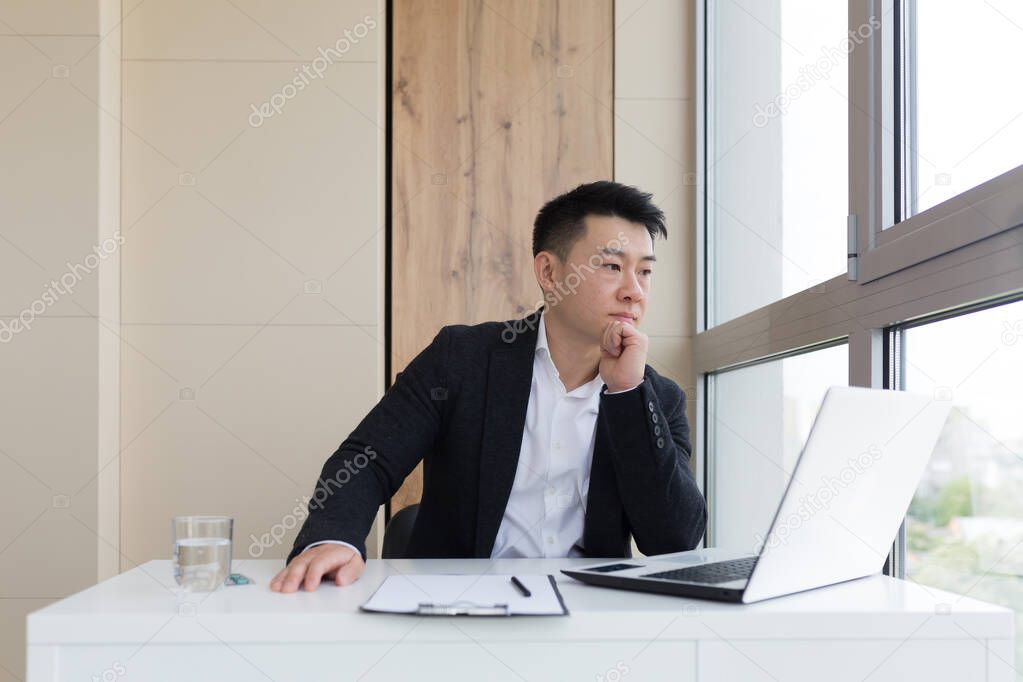 young asian office worker suffering from headache at work in the office drinks medicine pill with water. Sick man in a suit at the computer indoors with severe pain ache uses the drug in workplace