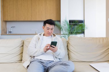 Asyalı adam evinde kanepede oturuyor, akıllı telefon kullanıyor, telefonda ciddi bir mesaj yazıyor.