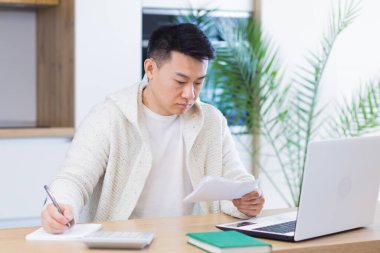 Evinde evrak işleriyle uğraşan genç Asyalı bir adam masa başında oturuyor, dizüstü bilgisayarda çalışıyor, faturaları ödemeye odaklanıyor.