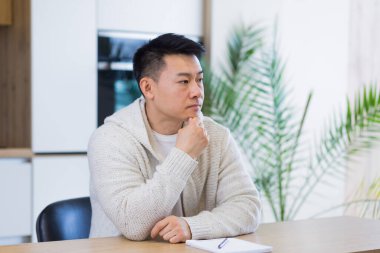 Düşünceli, ciddi, Asyalı genç bir adam başka tarafa bakıyor. Öğrenci yazar evde dizüstü bilgisayarla oturup düşüncelere dalmış, düşüncelere dalmış, finansal sorunları çözmeyi düşünüyor.