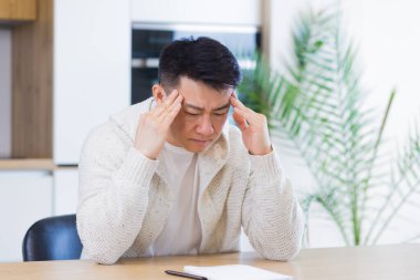 Düşünceli, ciddi, Asyalı genç bir adam başka tarafa bakıyor. Öğrenci yazar evde dizüstü bilgisayarla oturup düşüncelere dalmış, düşüncelere dalmış, finansal sorunları çözmeyi düşünüyor.