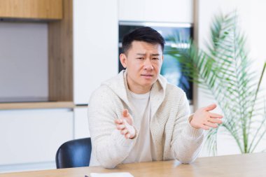 Asyalı adam evdeki ya da mutfaktaki video konuşmasında konuşuyor. Erkek, günlük giysiler içinde kameraya bakıyor, toplantı yapıyor ya da uzaktan konferans veriyor. Portre öğretmeni. Ders eğitimi. Web kamerası görünümü