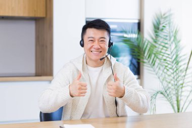 Asyalı bir adam, video odasında kulaklıkla konuşuyor. Sıradan giyinmiş bir erkek kameraya, toplantıya ya da uzaktan konferansa bakıyor. Portre öğrencisi ya da öğretmen eğitimi. Web kamerası görünümü