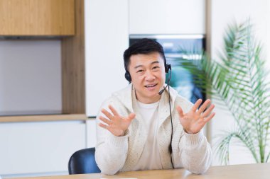 Asyalı bir adam, video odasında kulaklıkla konuşuyor. Sıradan giyinmiş bir erkek kameraya, toplantıya ya da uzaktan konferansa bakıyor. Portre öğrencisi ya da öğretmen eğitimi. Web kamerası görünümü