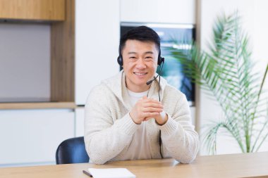 Asyalı bir adam, video odasında kulaklıkla konuşuyor. Sıradan giyinmiş bir erkek kameraya, toplantıya ya da uzaktan konferansa bakıyor. Portre öğrencisi ya da öğretmen eğitimi. Web kamerası görünümü