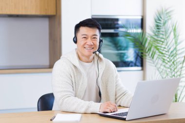 Kulaklıklı genç Asyalı adam kameraya bakıyor ve mutfakta oturmuş gülümsüyor ve dizüstü bilgisayarla çalışıyor.