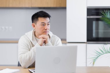 Düşünceli, ciddi, Asyalı genç bir adam başka tarafa bakıyor. Öğrenci yazar evde dizüstü bilgisayarla oturup düşüncelere dalmış, düşüncelere dalmış, finansal sorunları çözmeyi düşünüyor.