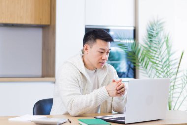 Evde dizüstü bilgisayarla çalışan Asyalı genç adam mutfaktaki masada oturuyor.