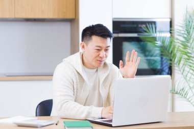 Genç Asyalı adam evde, oturma odasında ya da mutfakta oturuyor ve internetten iletişim kuruyor. Dizüstü bilgisayarın web kamerasında erkek el sallıyor. Video 'ya öğrenme mesafesi. Eğitim, konferans veya E-öğrenim toplantısı