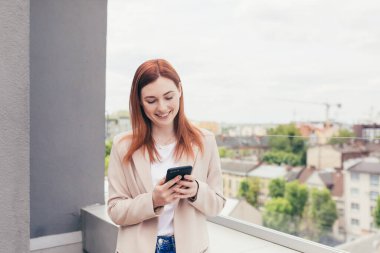 Genç ve güzel bir kadın telefonda yazışıyordu. Gülümsüyordu ve balkonda takım elbise giyiyordu.