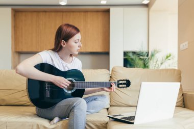 Genç kızıl saçlı kız video derslerinin yardımıyla gitar çalmayı öğreniyor. Bayan kadın boş vakitlerinde kanepede dizüstü bilgisayarla oturup online müzik aleti eğitimi alıyor. Hobiler mesafesi