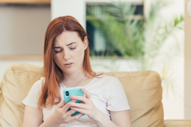 Evde oturan genç bir kadın, üzgün ve depresif. Kanepede oturan telefondan kötü haberler okuyor.