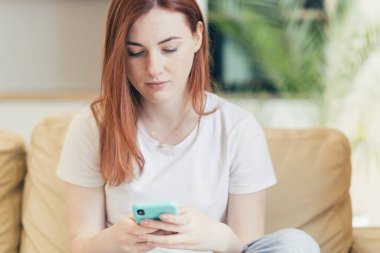 Evde oturan genç bir kadın, üzgün ve depresif. Kanepede oturan telefondan kötü haberler okuyor.