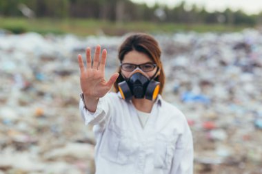 Çöp sahalarında korunan bir solunum giysisi giymiş bir kadın, elele tutuşmuş, gezegenin kirliliğini durdurmaya çalışıyor, elleriyle bir dur işareti gösteriyor.