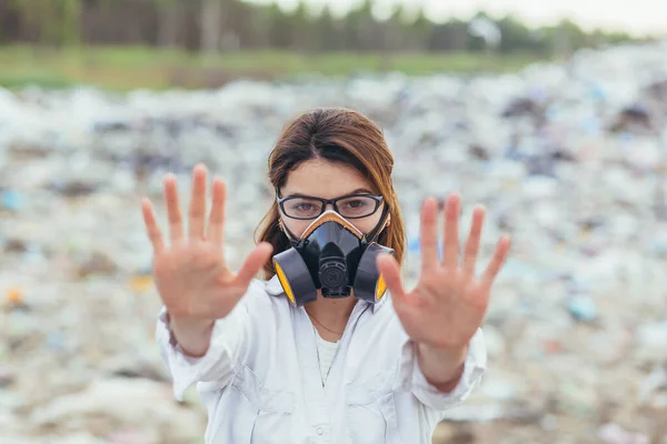 A woman in a protected respiratory suit at landfills, holding hands in front, trying to stop the pollution of the planet, shows a stop sign with her hands