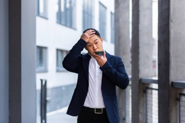 Sinirli ve ciddi Asyalı başarılı iş adamı telefon kullanarak çalışanlara bilgi veriyor, ofis dışında konuşuyor