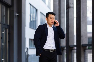 Sinirli ve ciddi Asyalı başarılı iş adamı telefon kullanarak çalışanlara bilgi veriyor, ofis dışında konuşuyor