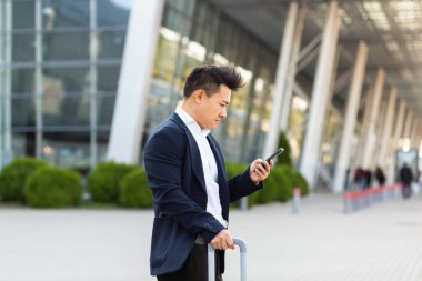 İş adamı bir uygulama ve cep telefonu kullanarak taksi çağırmaya çalışıyor. Tren istasyonundaki büyük bavullu Asyalı adam.