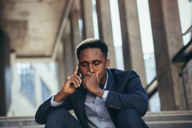 Depresyonda olan Afrikalı Amerikalı iş adamı cep telefonuyla konuşuyor. Ofis merdivenlerinde takım elbisesiyle oturuyor.
