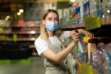 Tezgahtar kadın portresi, koruyucu maskeli kadın gülümseyen ve süpermarkette kameraya bakan. Dost canlısı bayan satıcı sıraların arasında mağazada duruyor. Çapraz kollu işçi