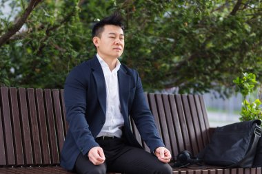 Asyalı işadamı stresi yatıştırmak için nefes alma egzersizleri yapıyor, iş elbisesiyle öğle yemeği arasında bankta oturuyor.