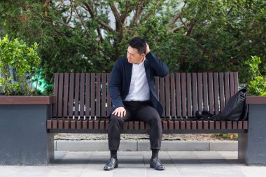 Asyalı işadamı işten sonra yorgun, yorgunluk ve baş ağrısı olan bir bankta oturuyor, stres ve panik atak geçiriyor.