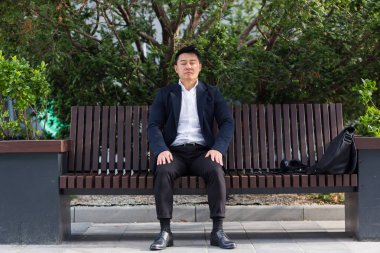 Asyalı işadamı işten sonra yorgun, yorgunluk ve baş ağrısı olan bir bankta oturuyor, stres ve panik atak geçiriyor.