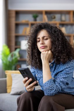 Young woman sitting on a sofa looking worried and disappointed while holding her mobile phone, reading unsettling news or a message with a concerned expression