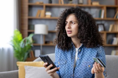 Woman with curly hair feeling stressed and confused, holding a credit card and smartphone, encountering an online payment error or fraud while shopping from home