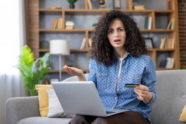 Woman feeling frustrated and confused, holding a credit card and looking at her laptop screen after encountering a problem with an online payment while shopping from her couch