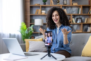 Woman with curly hair sitting on a sofa and recording a video blog with her smartphone on a tripod, gesturing actively while live streaming her content