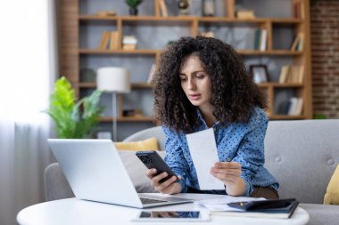 Woman reviewing household finances on laptop and phone, calculating bills and receipts in living room while managing budget, online payments and personal expenses