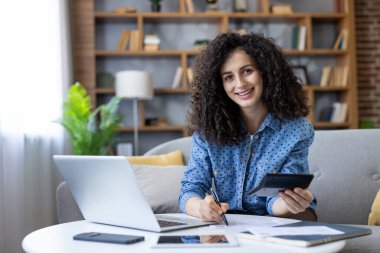 Young woman smiling, analyzing financial documents, calculating expenses using a laptop and calculator while working from a comfortable home office setup, managing her budget