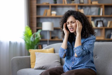 Woman experiencing intense migraine pain, sitting on a comfortable couch in her living room, placing hands on temples, finding temporary relief from discomfort, feeling unwell and needing rest