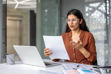 Stressed businesswoman in an office reviewing a financial document, feeling overwhelmed and confused by bad news or unexpected problems, with a laptop nearby