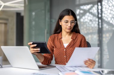 Indian businesswoman in modern office reviewing financial reports, using laptop and calculator while focused on budgets, accounting and investment documents at her desk