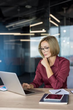Businesswoman with glasses collaborating and concentrating at her modern office workspace, using a laptop and other digital devices to manage her business tasks efficiently