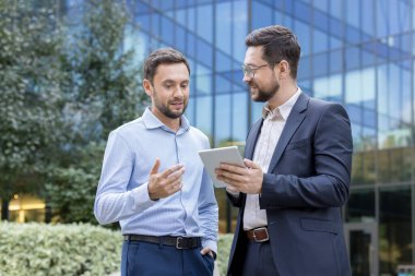 Two businessmen collaborate outdoors, reviewing ideas and data on a tablet in front of a modern glass office building, discussing strategy, planning and partnership