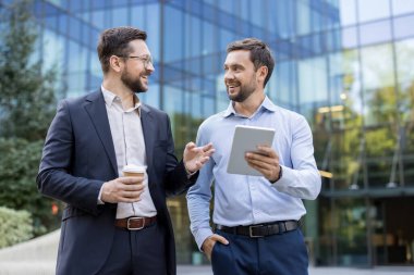Two cheerful businessmen stand by a modern office building, holding a tablet and coffee, smiling and chatting outdoors during a casual work break about strategy and collaboration